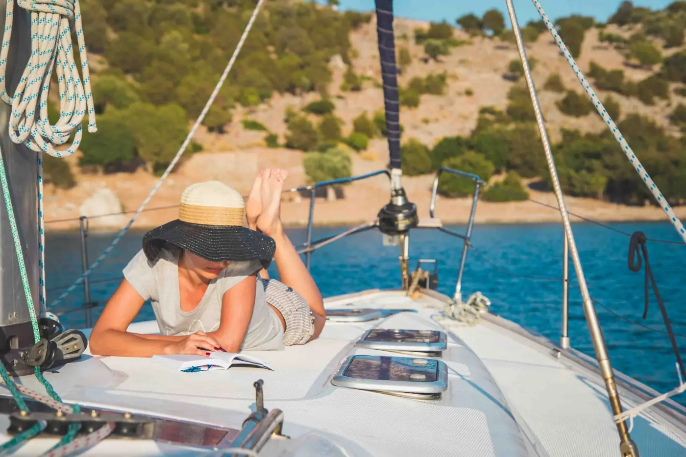 A woman reading a book on the deck of a sailboat anchored near the shore