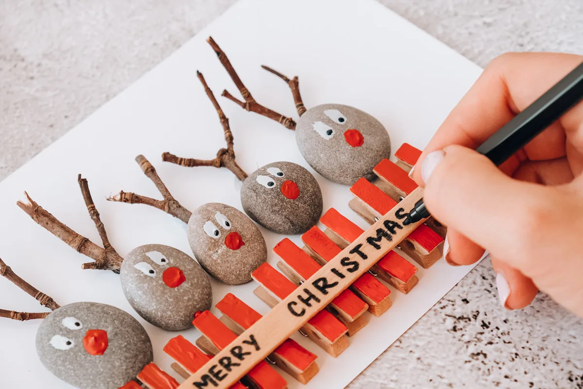 A hand writing Merry Christmas beside handmade pebble reindeer decorations